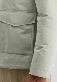 Lichtgrijze jas mouw en grote klepzak detail op beige broek, weergegeven aan de rechterzijde en arm van een persoon.