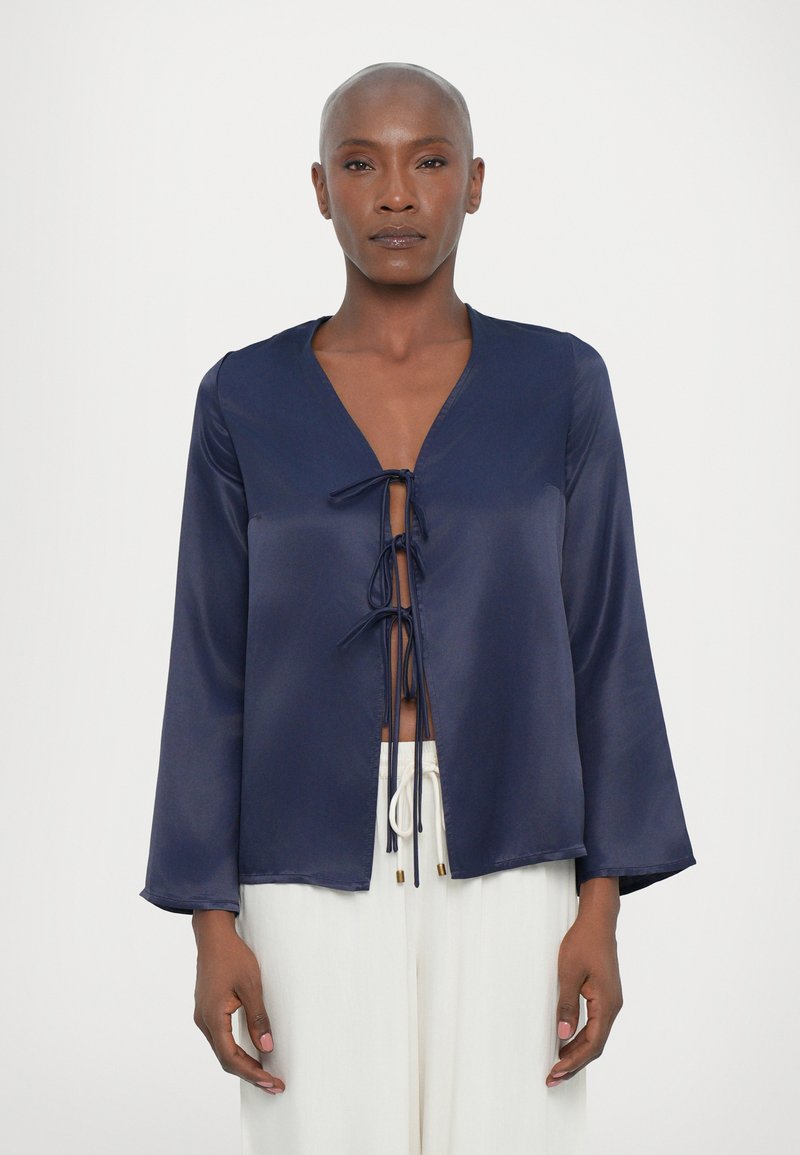 Blusa de satén azul marino de manga larga con escote en V y cierre con lazo en el frente. Presenta mangas anchas y un diseño corto. Textura suave.