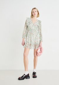 Cvetna mini obleka z dolgimi rokavi, v-izrezom, volančki; prevladujoča bela barva s roza in modrimi cvetlicami. Manekenka drži roza clutch, obuta v črne čevlje.