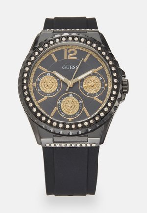 Montre Guess noire et dorée avec lunette ornée de cristaux, trois sous-cadrans pour la date, le jour et l’heure au format 24 heures, et un bracelet en silicone noir.
