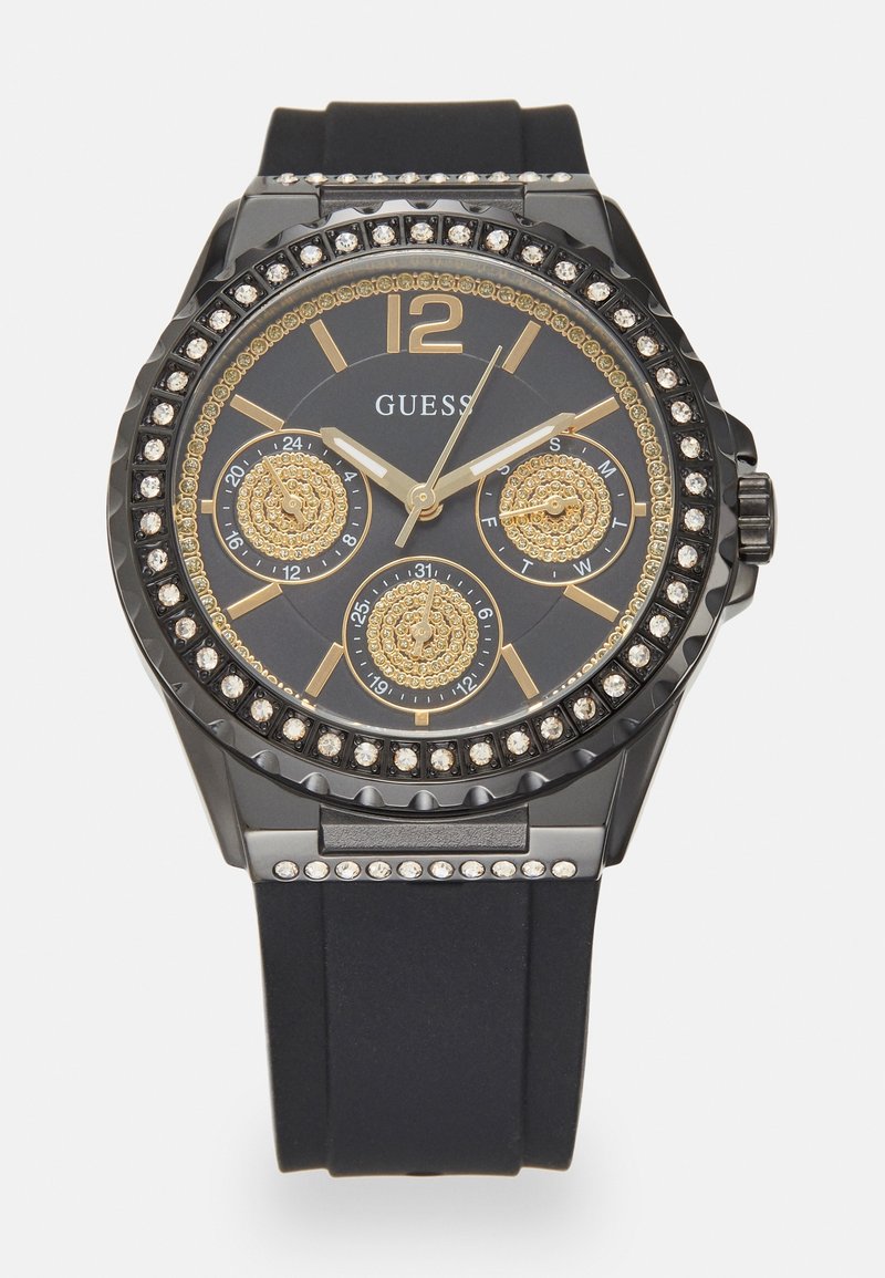 Montre Guess noire et dorée avec lunette ornée de cristaux, trois sous-cadrans pour la date, le jour et l’heure au format 24 heures, et un bracelet en silicone noir.