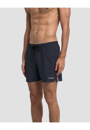 Mann trägt schwarze Badeshorts mit blauen und orangefarbenen Seitenstreifen und weißem Sundek-Logo am linken Bein, steht vor einem einfarbigen Hintergrund.