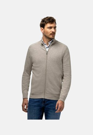 Man wearing light beige zip-up sweater over light blue shirt and blue jeans, looking to the side with neutral expression.