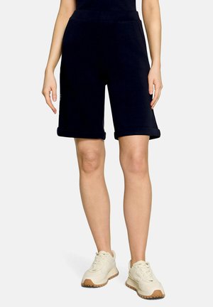 Personne portant un short noir jusqu'aux genoux et des baskets blanches, debout devant un fond clair uni.