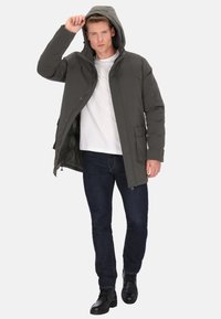 Jachetă izolată verde închis cu glugă, închideri cu fermoar, buzunare frontale și textură netedă, purtată peste un tricou alb și blugi închiși la culoare.