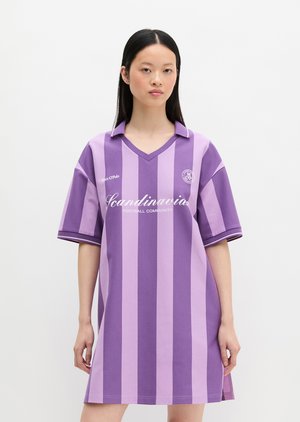 Tânără purtând o rochie cămașă largă cu dungi mov și lavandă, cu textul „Scandinavia Football Community” pe față.