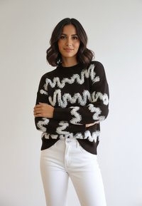 Kobieta o ciemnych włosach do ramion, ubrane w czarny sweter z białymi falistymi wzorami i białe spodnie, stoi ze skrzyżowanymi ramionami.