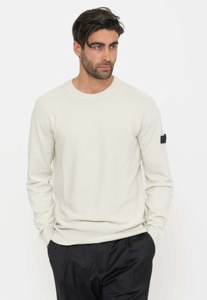 Uomo con capelli scuri e barba che indossa un maglione color crema chiaro e pantaloni neri in piedi con le mani nelle tasche, guardando di lato.