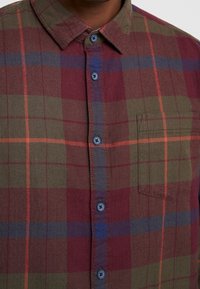 Camicia a caramelli nei toni del verde, bordeaux e blu, con colletto button-down, bottoni blu e taschino sul petto con orlo cucito.