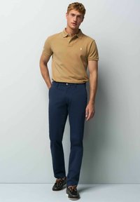 Beige polo shirt with a collar, navy pants, and dark shoes. Polo features a small logo on the chest. Textured cotton fabric.