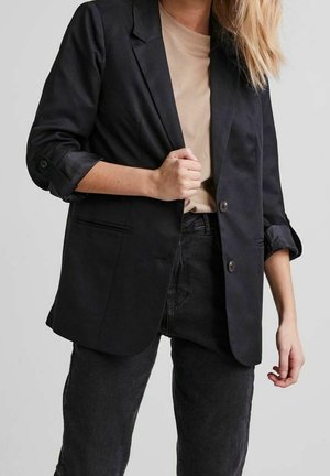 Černý blazer s jedním řadou knoflíků, s klopami, knoflíkovými rukávy a dvěma předními kapsami. Nositý přes béžový top a černé džíny.