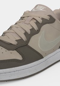 Zbliżenie beżowo-ciemnobrązowego buta sportowego Nike z białą podeszwą i sznurówkami, na boku widoczny jest logo Nike swoosh oraz oznaczony język.