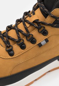 Bota de senderismo de cuero marrón con cordones negros, ojales de plástico, una mediasuela blanca y una suela de goma negra texturizada.