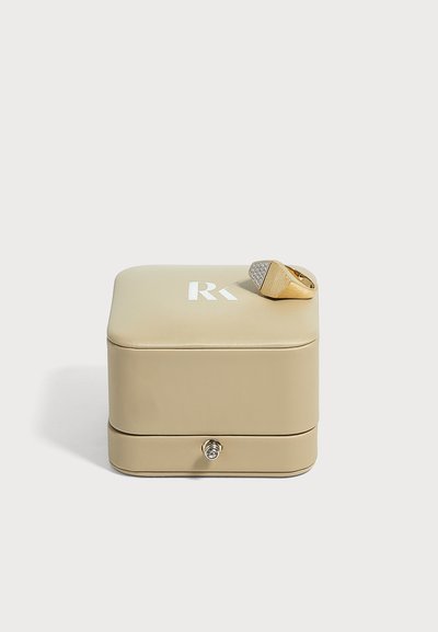 Boîte à bijoux beige avec un logo RK argenté sur le dessus et une bague en or avec une face triangulaire sertie de diamants posée dessus.