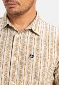 Camisa de manga corta de color beige con un patrón que presenta rayas verticales y diseños florales. Incluye un pequeño parche negro con el logotipo y un cierre frontal de botones.
