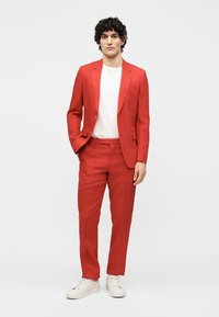 Άνδρας με σγουρά μαλλιά, που φοράει κόκκινο κοστούμι, λευκό μπλουζάκι και λευκά αθλητικά παπούτσια, στέκεται με το ένα χέρι στην τσέπη μπροστά σε απλό φόντο.