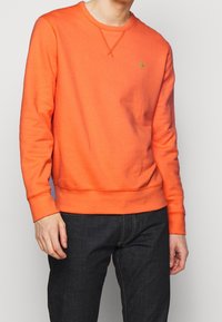 Orange sweatshirt med rund halsringning, ribbade ärmar och fåll. Har en liten grön logotyp på bröstet. Mjuk textur och avslappnad passform.