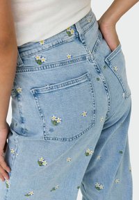 Lichtblauwe denim jeans met verspreide witte madeliefjes borduursels. Losse pasvorm met zakken en contrasterende stiksels.
