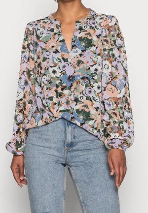 Kobieta ubrana w długorękawową, kwiatową bluzkę w odcieniach fioletu, zieleni i brzoskwini, zestawioną z jasnoniebieskimi, wysokim stanem jeansami.