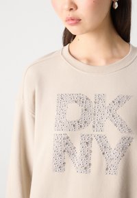 Bežinis džemperis su tekstūru ir permatomu DKNY logotipu, puoštas cirkoniu, apvaliu iškirpimu ir laisvu siluetu.