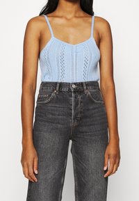 Top de punto azul claro con tirantes finos, que presenta un patrón texturizado. Combinado con unos vaqueros de mezclilla negros de cintura alta.