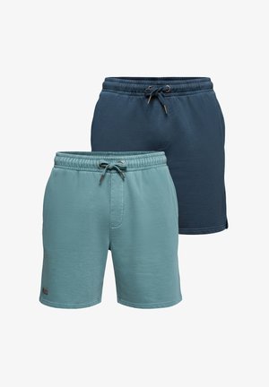Mann trägt weiße Kappe, lockeres weißes T-Shirt, türkisfarbene Shorts und weiße Sneakers, posiert mit einer Hand in der Tasche und der anderen, die seine Kappe berührt.