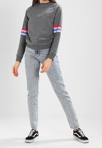 Grijze sweatshirt met rode, blauwe en paarse strepen op de mouwen. Gecombineerd met lichtblauwe, hooggetailleerde spijkerbroek en zwarte sneakers.