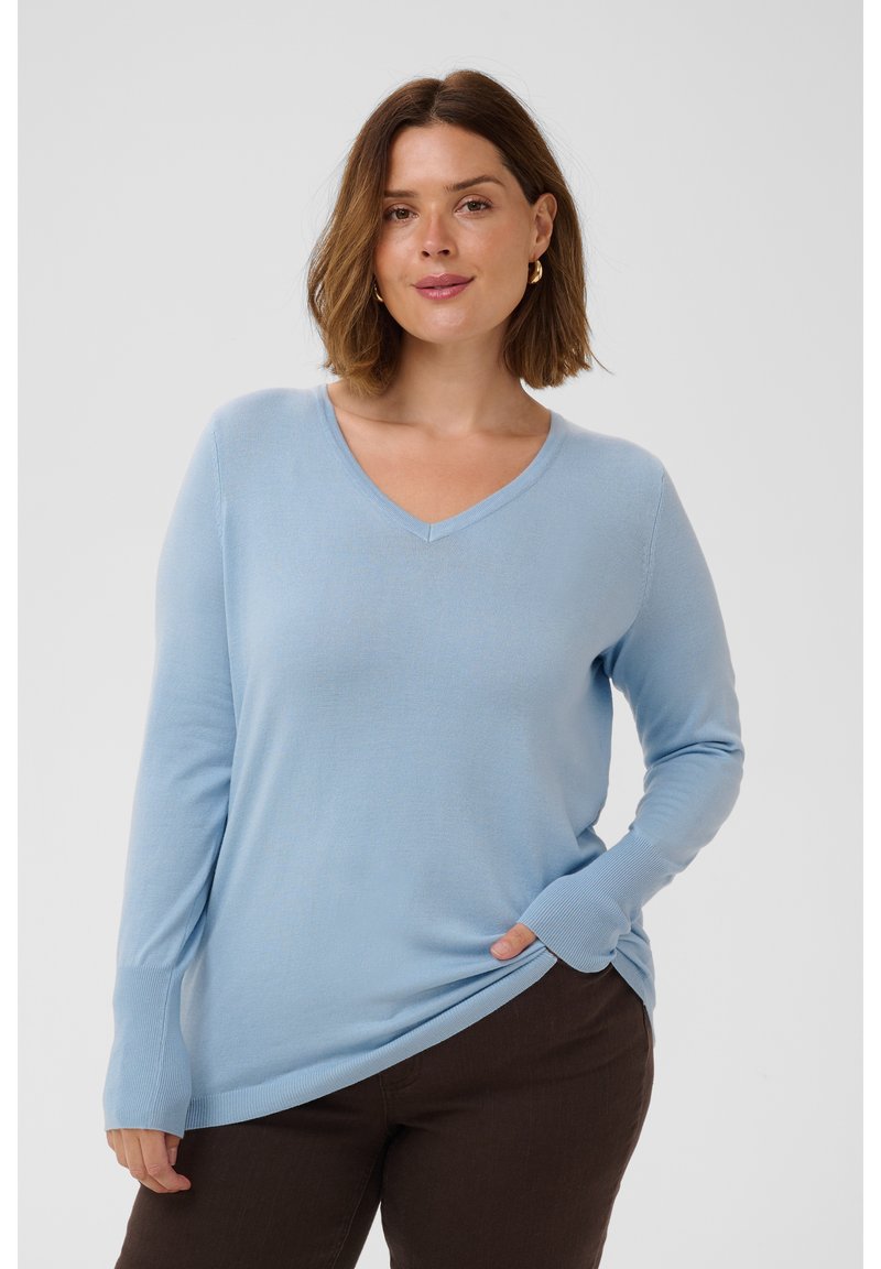 Femme aux cheveux courts bruns, portant des boucles d’oreilles créoles dorées, un pull bleu clair à col en V et un pantalon foncé sur un fond uni.