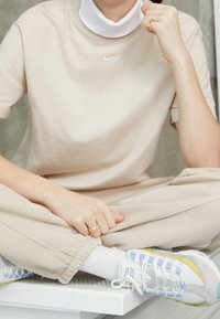 Beige bomulls-T-shirt med en vit Nike-logotyp, lager på lager över en vit höghalsskjorta. Beiga joggingsbyxor och pastellfärgade sneakers fulländar looken.