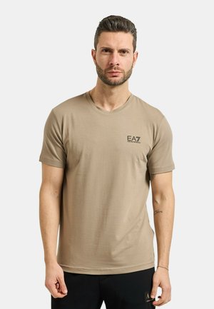 Uomo con capelli corti e scuri e barba che indossa una t-shirt beige EA7 Emporio Armani e pantaloni neri su uno sfondo chiaro e semplice.