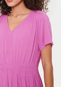 Růžové šaty s krátkým rukávem a véčkovým výstřihem, vyrobené z texturované tkaniny; mají nařasený pas a splývavou sukni. Vypadají pomačkaně.
