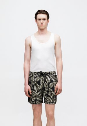 Young man wearing white sleeveless tank top and black swim shorts with beige tropical leaf print, standing against plain light background.