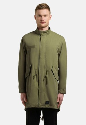 Mężczyzna w oliwkowo-zielonym długim płaszczu parka z wysokim kołnierzem, kieszeniami z patkami oraz ściągaczami z przodu i na dole, stojący na jednolitym tle.