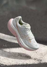 Άσπρο και ροζ αθλητικό παπούτσι με επάνω μέρος από δίχτυ και κορδόνια, αιωρούμενο στον αέρα πάνω από μια ασφαλτωμένη επιφάνεια με λευκές οδικές σημάνσεις.