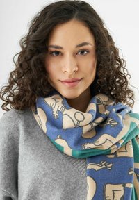Écharpe tricotée présentant un motif d'éléphant bleu et beige, avec une texture douce et un tissu fluide, portée sur un pull gris.