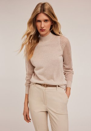 Suéter de punto beige claro con cuello alto, con acentos de canalé en las mangas. Combinado con pantalones color crema y un cinturón decorativo.
