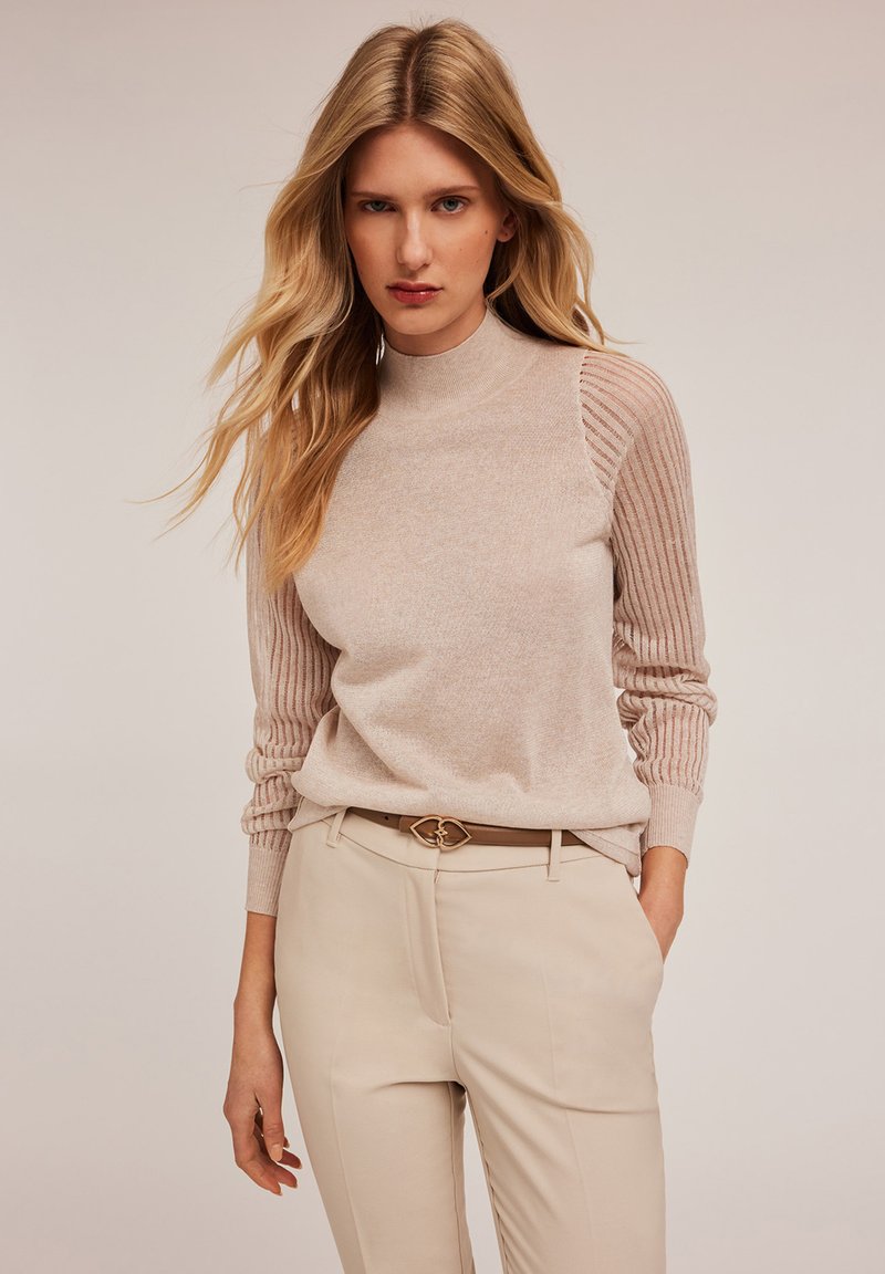 Hellbeige Strickpullover mit hochgeschlossenem Kragen, der gerippte Akzente an den Ärmeln aufweist. Kombiniert mit cremefarbenen Hosen und einem dekorativen Gürtel.