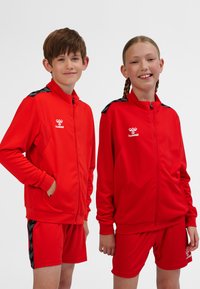 Red sports tracksuits made of textured fabric. Jackets feature zippers, side pockets, and contrasting black accents on shoulders.