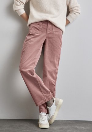 Personne portant un pantalon en velours côtelé rose clair, un pull crème, des chaussettes grises et des baskets blanches, debout contre un mur gris.
