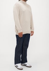 Pull à col roulé beige clair avec coupe décontractée, associé à un jean droit bleu foncé et à des baskets grises avec des rayures noires.