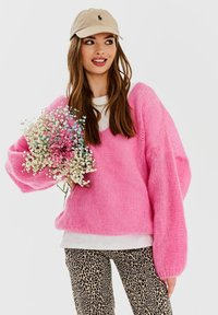 Różowy, oversize'owy sweter z dzianiny o teksturowanych rękawach, noszony na białej koszuli, w połączeniu z spodniami we wzór panterki. Modelka trzyma bukiet kwiatów.