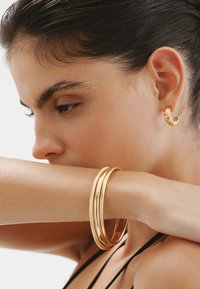 Pendiente de aro en tono dorado con diseño texturizado; pulseras de oro apiladas con un acabado liso y estriado sobre un tono de piel claro.