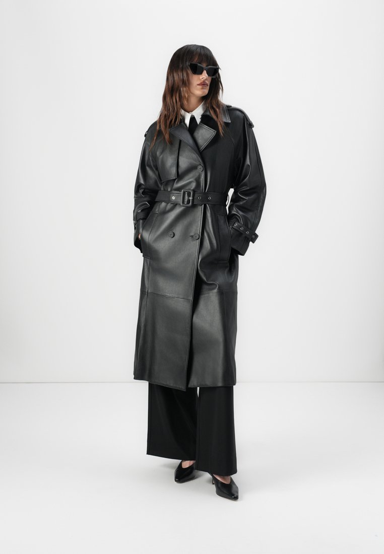 Manteau trench en cuir noir avec une taille ceinturée, un devant croisé et des poignets larges, associé à un pantalon large noir et des talons.