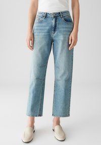 Vrouw in lichtblauwe rechte jeans met gestikte kniedetails en off-white instappers tegen een effen achtergrond.