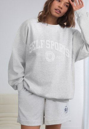 Kobieta w jasnoszarej, oversize'owej bluzie z nadrukiem "SELF SPORTS" oraz pasujących szortach stoi w pomieszczeniu, trzymając rękę przy czole.