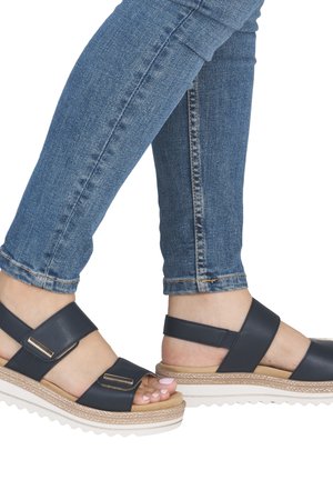 Navy blue platform sandals with two wide straps, gold accents, and a textured beige sole. Paired with slim-fit blue jeans.