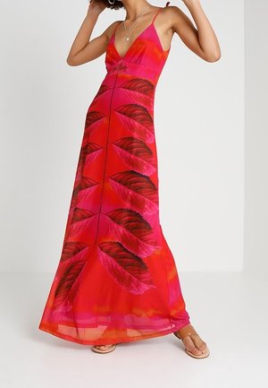 Mujer con vestido largo rojo sin mangas con patrones de hojas en rosa oscuro y negro y tirantes finos, de pie con un brazo levantado.