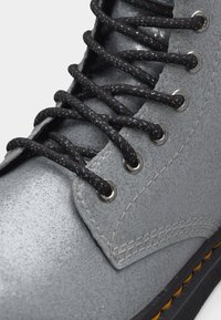 Κοντινή λήψη γκρι δερμάτινης μπότας με κορδόνια μαύρου χρώματος με λευκές βούλες, περασμένα από μεταλλικά κορδονότρυπες, και κίτρινο ράφωμα στη μαύρη σόλα.