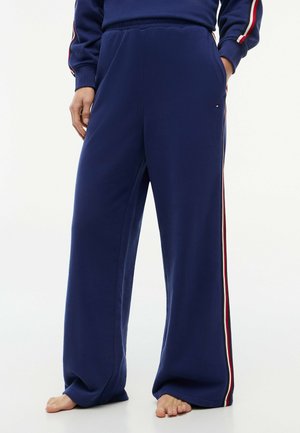Persona con pantalones anchos azul marino con franjas laterales rojas, blancas y azules, de pie descalza con las manos en los bolsillos.