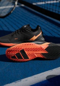 Μαύρο αθλητικό παπούτσι με πορτοκαλί λεπτομέρειες, που διαθέτει υφή στην επάνω επιφάνεια, ανθεκτική σόλα με σχέδιο ψαροκόκαλου και ενισχυμένη φτέρνα για υποστήριξη.